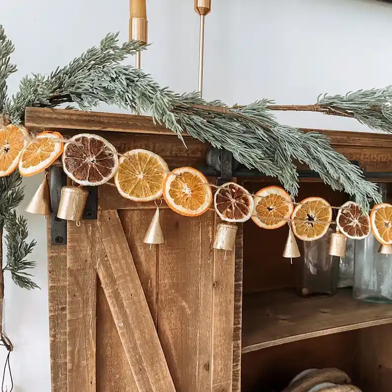 Home decor ideas, a DIY orange garland on cabinet with brass bells and festive greenery.